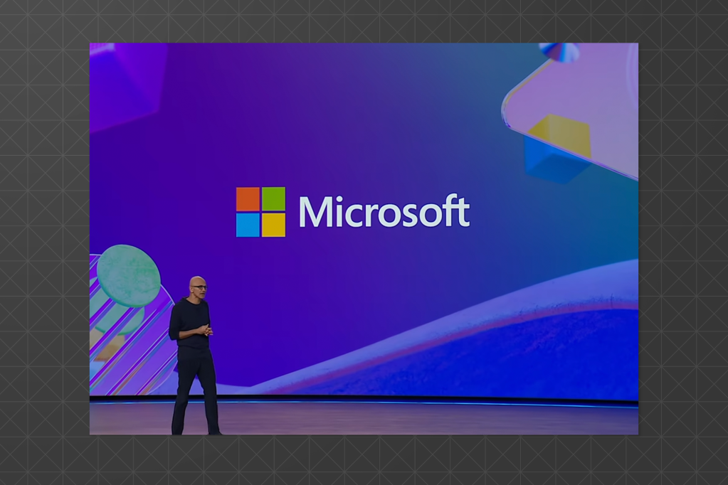 Satya Nadella on stage at a Microsoft event, with a large Microsoft logo on a purple background.
