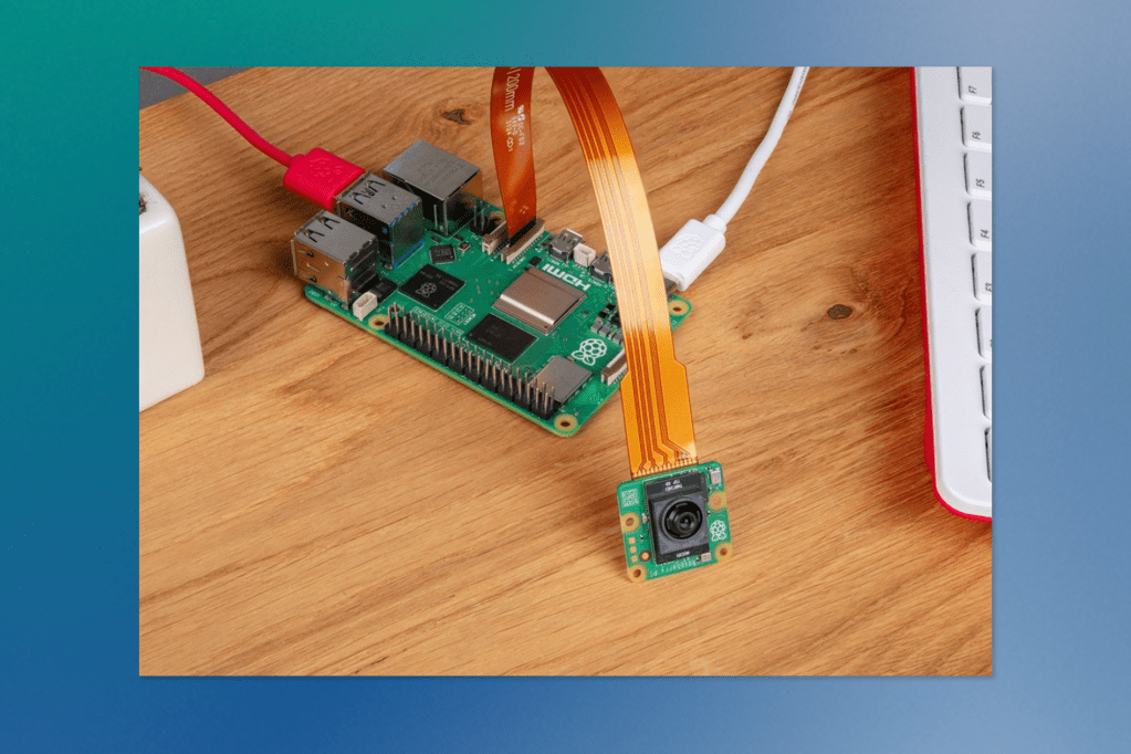 A Raspberry Pi board is shown connected to a camera module via a ribbon cable. The board is plugged in and resting on a wooden desk beside a white keyboard, demonstrating a typical DIY electronics setup.