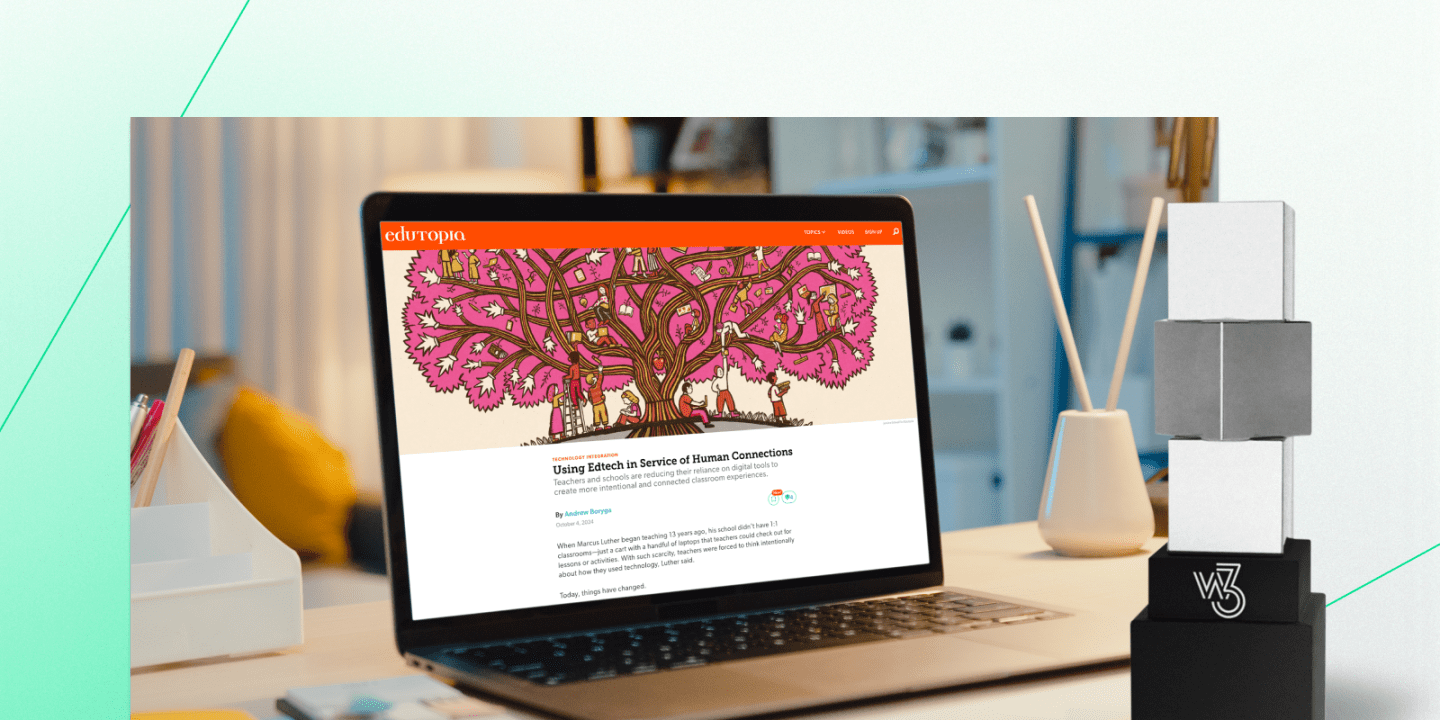 Person sitting at a desk viewing the Edutopia website on a laptop, displaying an article titled "Using Edtech in Service of Human Connections." The screen features a vibrant illustration of a tree filled with people. The workspace includes notebooks, a pen holder, and a glass bottle, set in a softly lit room.