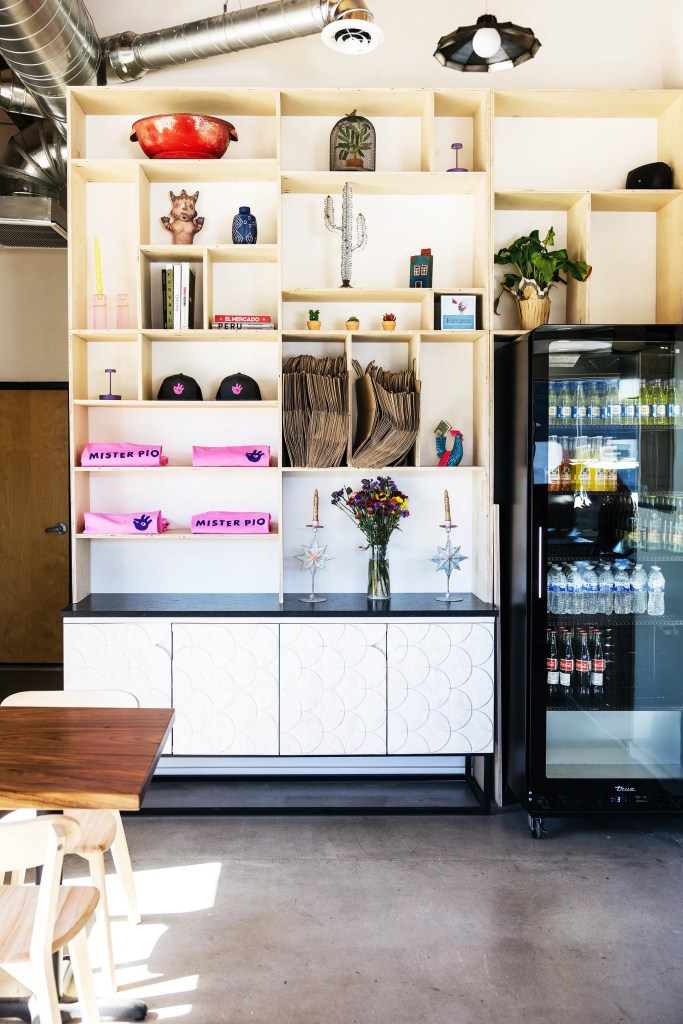 A modern display shelf filled with decorative items, plants, and books, next to a sleek refrigerator in a bright café setting. The wooden table in the foreground adds to the inviting atmosphere.