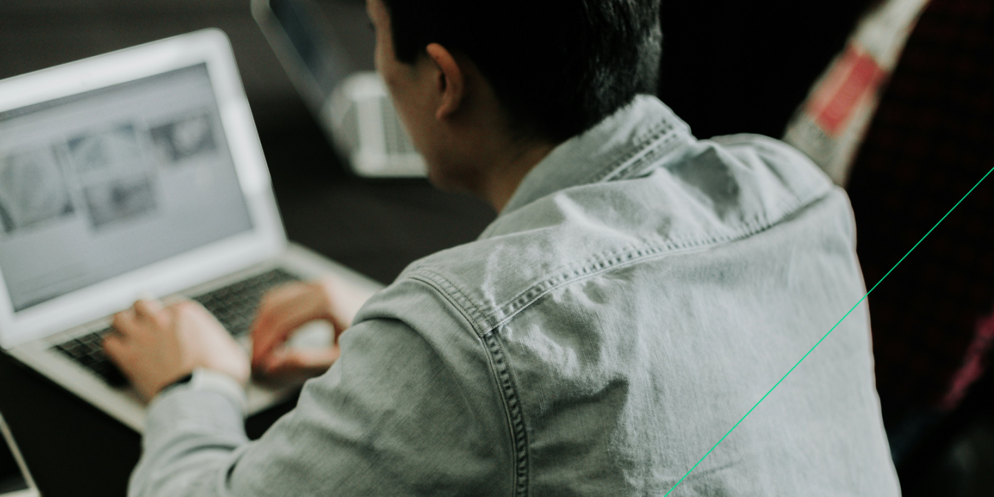 Person wearing a light denim shirt typing on a laptop, with another laptop and individual visible in the blurred background, suggesting a collaborative or work-focused environment.