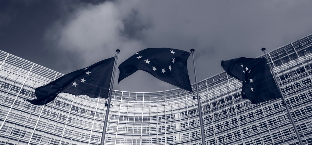Three European Union flags waving in front of a modern glass-fronted EU building under a cloudy sky, symbolizing EU governance and legislation.