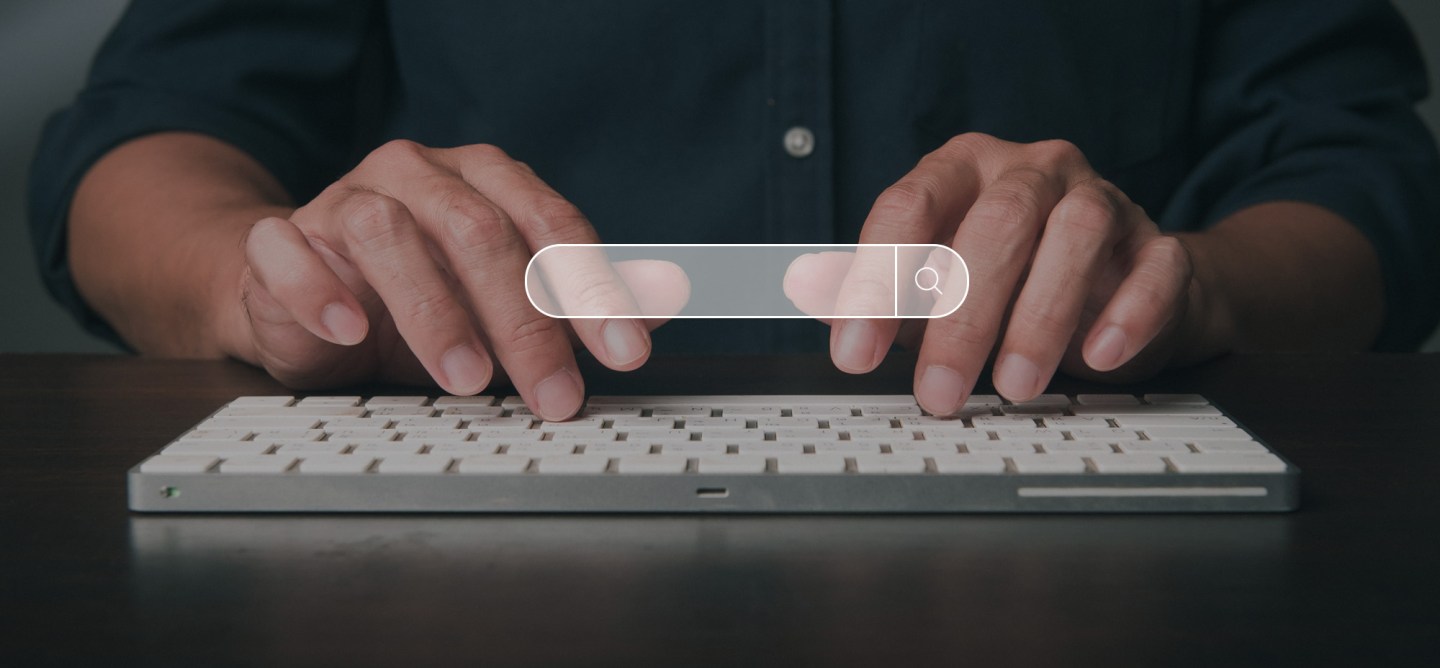 Person typing on a white keyboard with a floating digital search bar graphic overlayed above their hands, symbolizing online search or data entry.