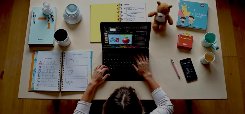Overhead view of a person using a laptop to design children's educational content, surrounded by notebooks, flashcards, colouring books, a schedule, and a teddy bear on a tidy desk.