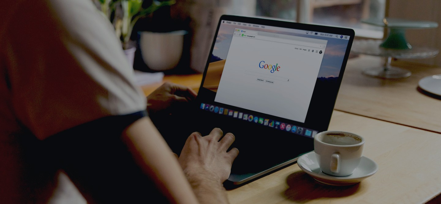 Person using a laptop at a wooden table with the Google homepage open on the screen. A cup of espresso sits beside the laptop. The setting appears cozy and softly lit, suggesting a café or home office environment, with blurred background elements like plants and glassware.