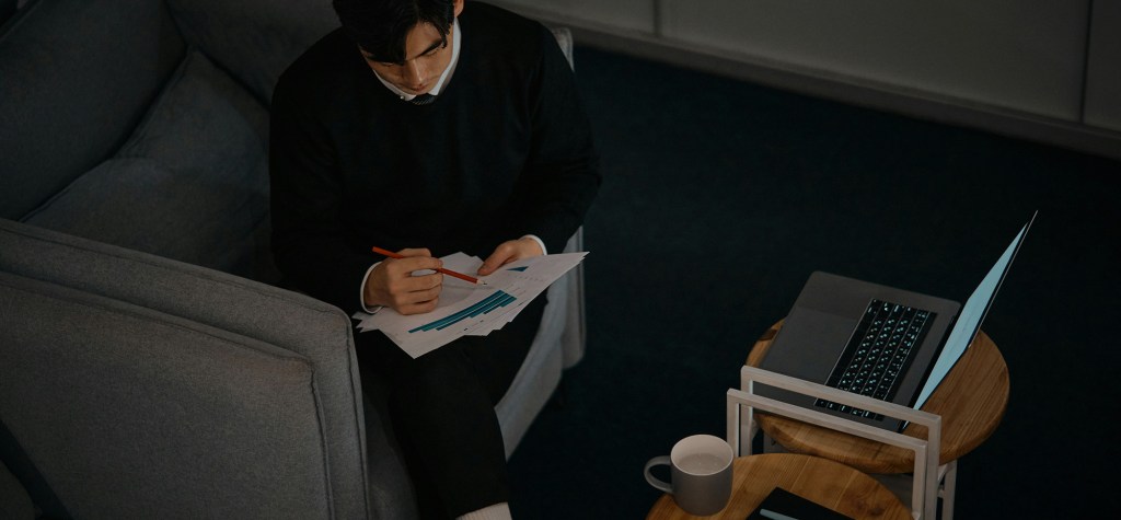 A man sits on a grey couch reviewing printed bar charts with a red pencil, while a laptop and coffee mug rest on nested wooden tables nearby.