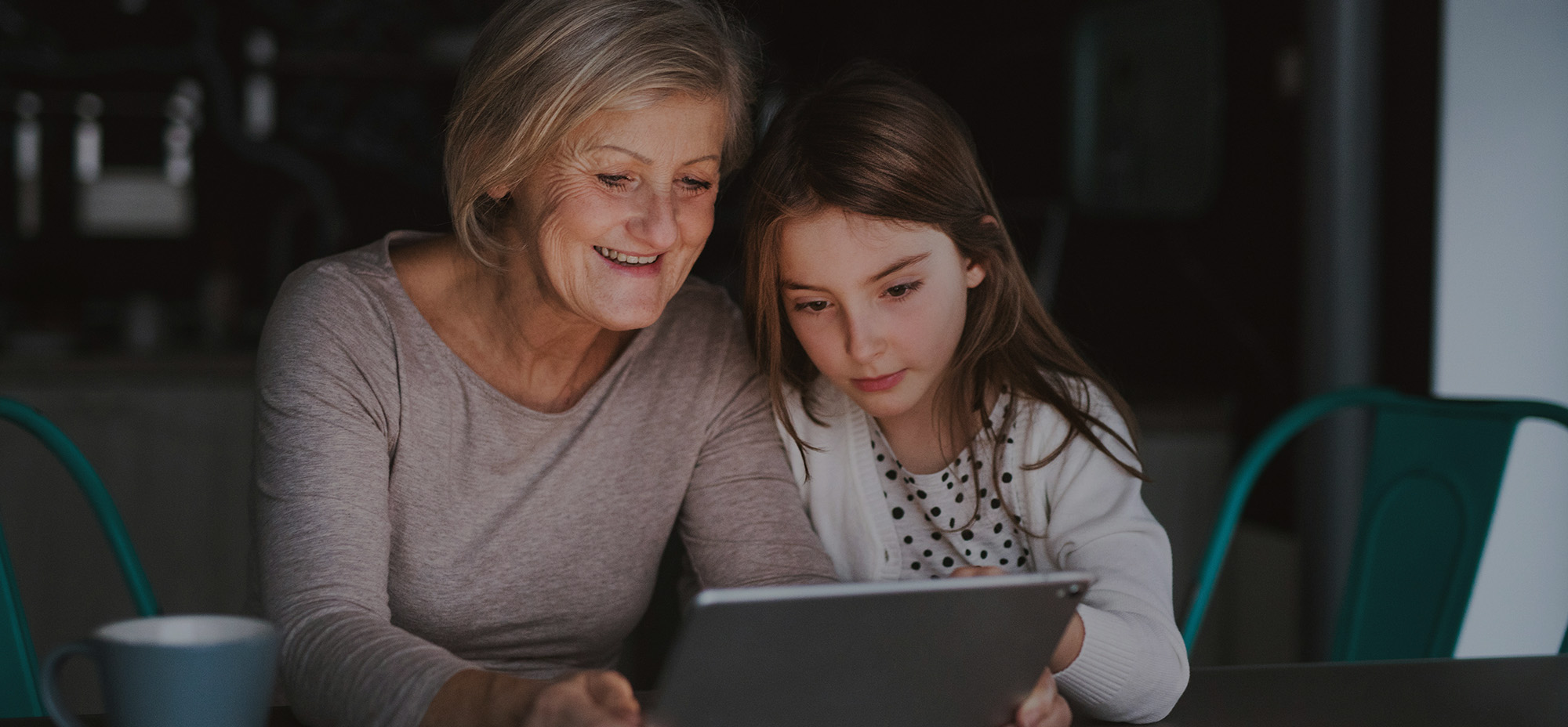 An elderly woman and a young girl sit closely together at a table, smiling as they look at a tablet screen. The scene suggests intergenerational learning or sharing digital experiences, with a coffee mug visible in the foreground.