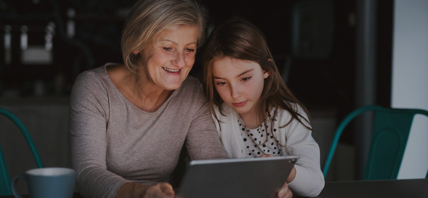 An elderly woman and a young girl sit closely together at a table, smiling as they look at a tablet screen. The scene suggests intergenerational learning or sharing digital experiences, with a coffee mug visible in the foreground.