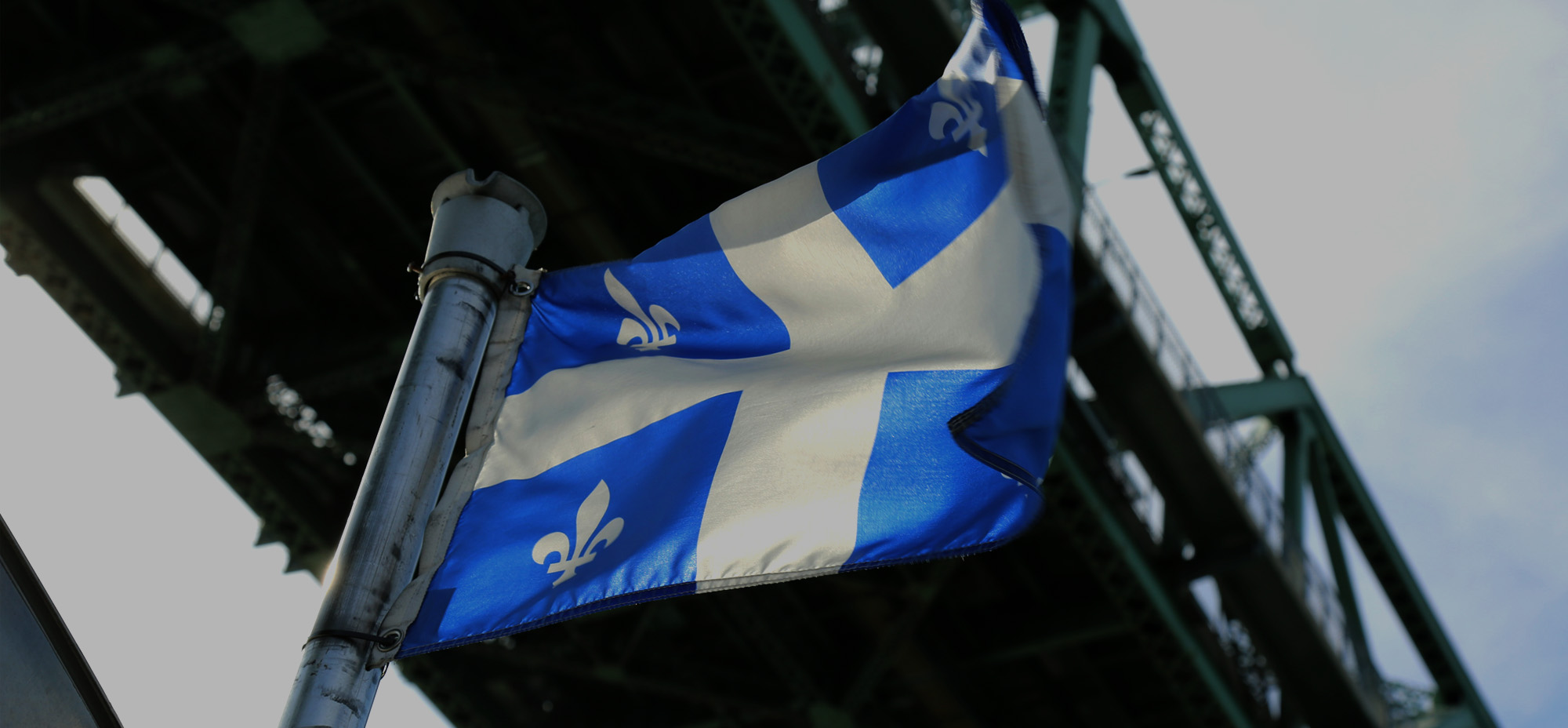 A close-up of the Quebec flag, known as the Fleurdelisé, flying in the wind under a steel bridge structure. The blue and white flag features four white fleurs-de-lis and a white cross on a blue field, symbolizing French heritage.