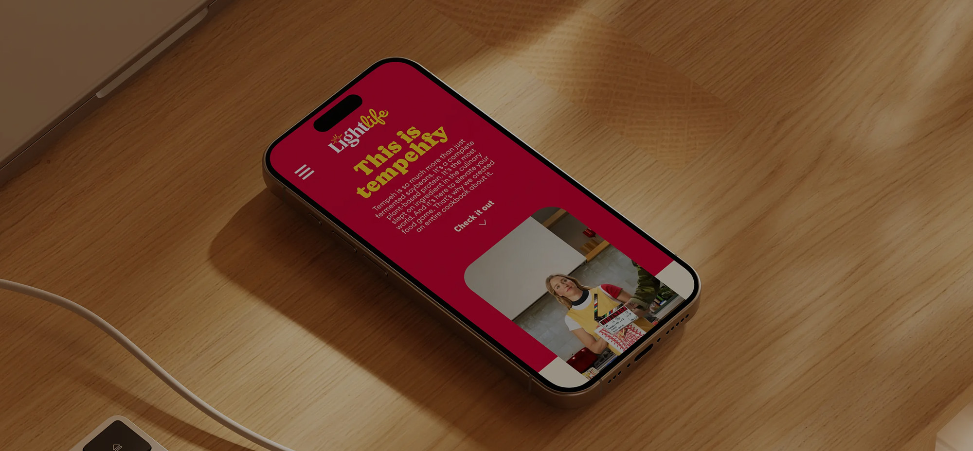 A smartphone resting on a wooden desk displays the Lightlife website with bold text reading “This is tempehfy” in yellow on a red background. A woman is featured below, promoting plant-based protein products.