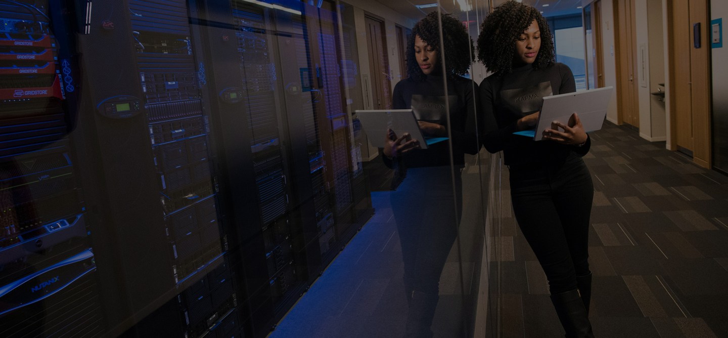 Woman in a black outfit holding a laptop while walking through a server room with data racks reflected on the glass wall.