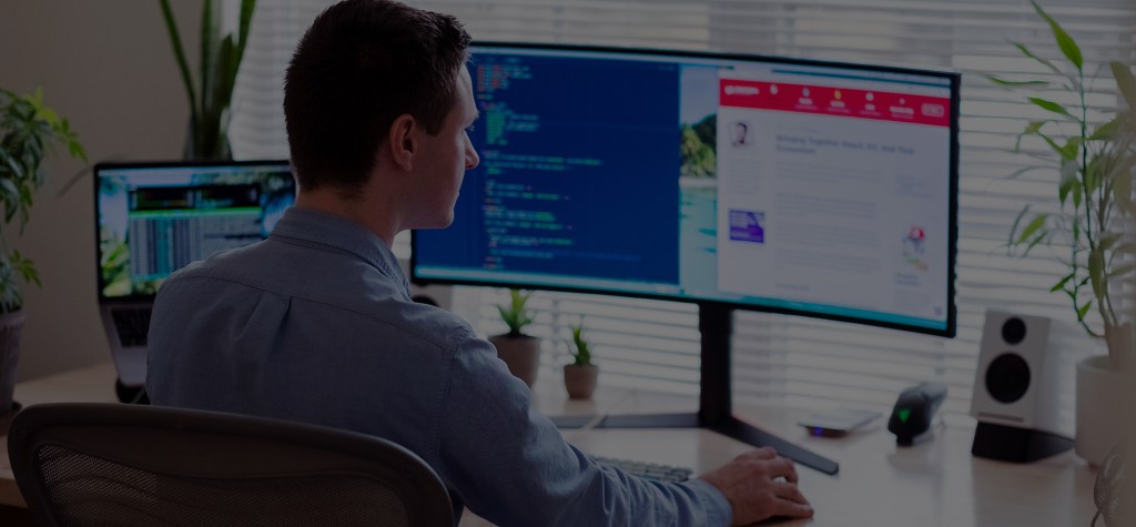 Man sitting at a desk with dual widescreen monitors, coding and reading content in a home office setup with plants and blinds.