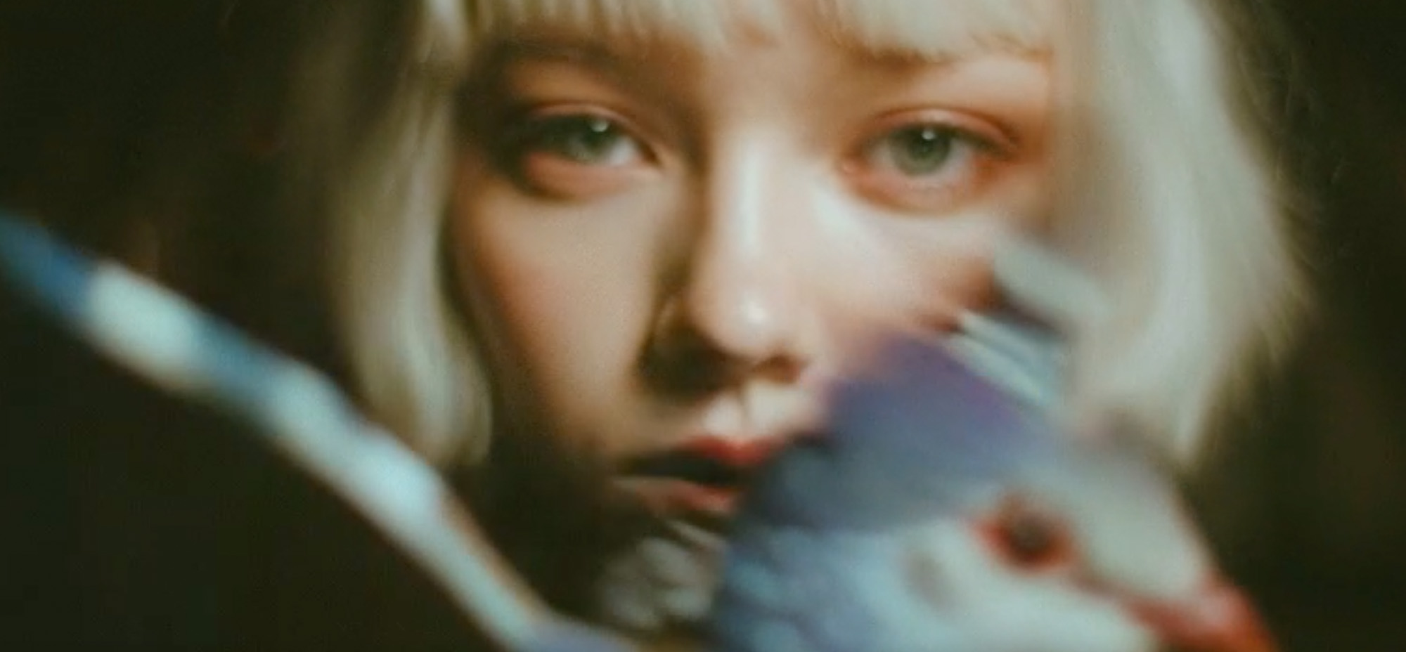 Close-up of a girl with platinum blonde hair looking directly at the camera, partially obscured by a bird in soft focus.