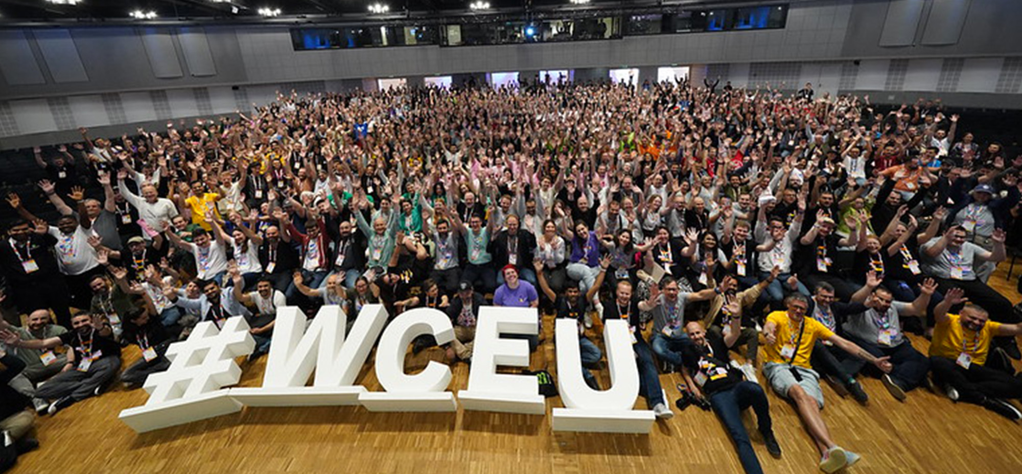 Large group photo of WordCamp Europe 2025 attendees celebrating in the Congress Center Basel, with hundreds of participants waving and cheering behind large white #WCEU letters on the wooden stage floor