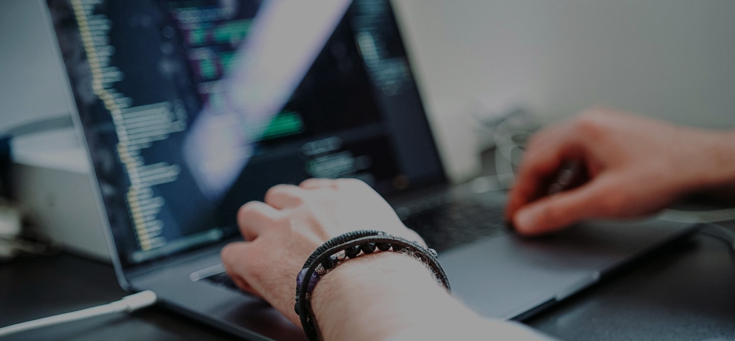 A person’s hands working on a laptop, with visible code on the screen. The image focuses on the act of programming or software development, with a bracelet visible on the wrist.
