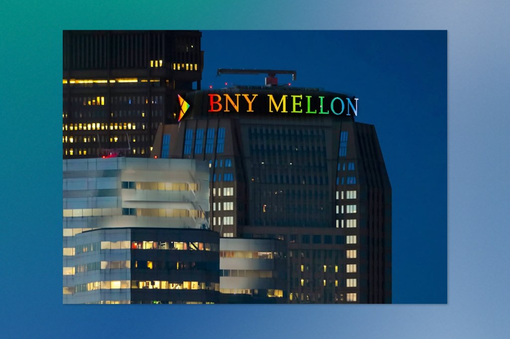 Night view of a tall office building with the BNY Mellon logo illuminated in rainbow colours on the top. The background includes other high-rise buildings with lit windows, creating a city skyline at dusk.