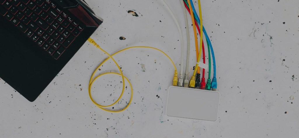 A flat lay image of a black laptop connected to a white hub via a yellow ethernet cable, with multiple coloured cables also plugged in, symbolizing digital connectivity and protocol integration.