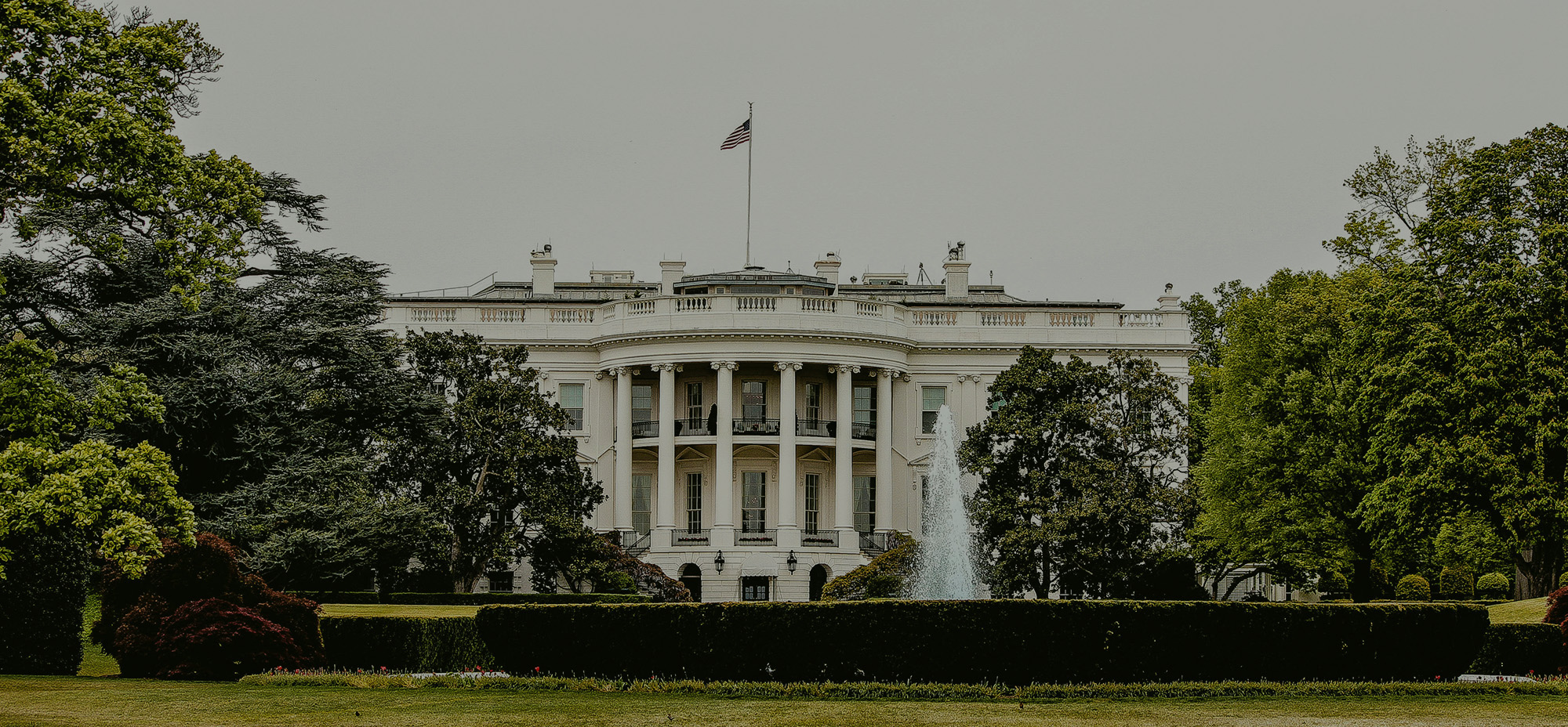 Front view of the White House in Washington, D.C., surrounded by lush green trees and manicured gardens. The U.S. flag is flying atop the historic building under an overcast sky.
