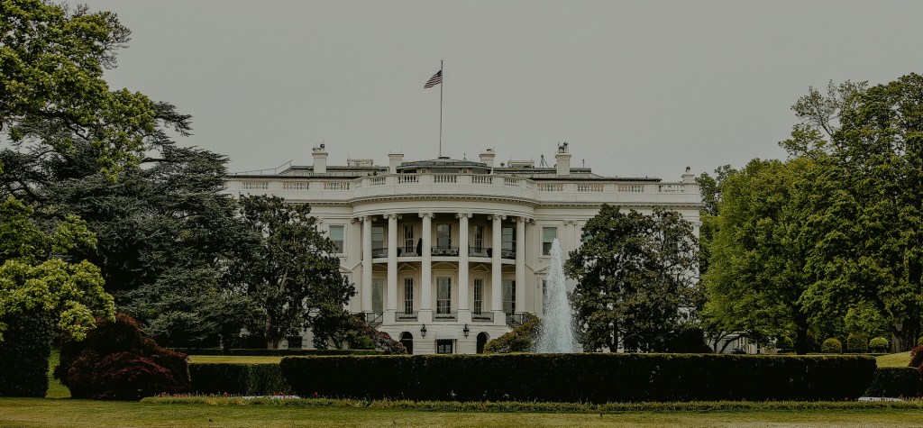 Front view of the White House in Washington, D.C., surrounded by lush green trees and manicured gardens. The U.S. flag is flying atop the historic building under an overcast sky.