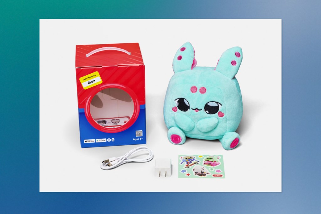 A bright teal plush toy named “Grem” with pink accents, displayed next to its packaging box, USB charging cable, wall adapter, and a sheet of colourful character stickers. The box includes branding for the Curio interactive AI toy.
