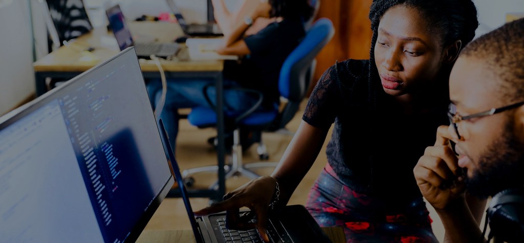 Two people collaborating at a desk, with one pointing at a laptop screen displaying code in a shared workspace.