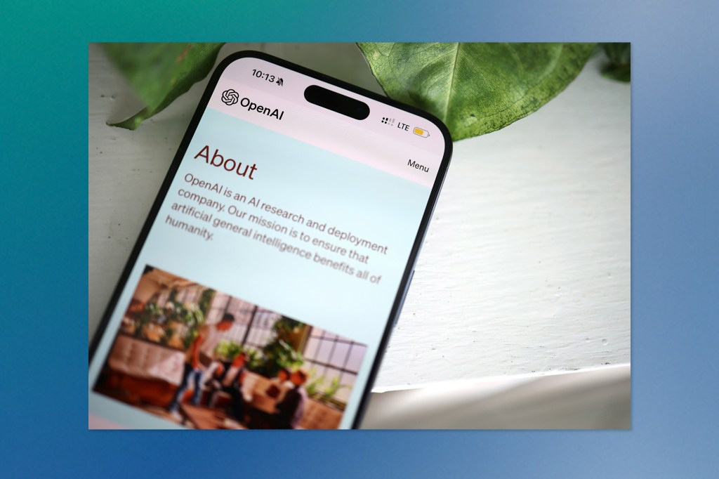 Smartphone screen displaying the OpenAI “About” page, with text explaining the company’s mission and a blurred photo of people in an office setting in the background.