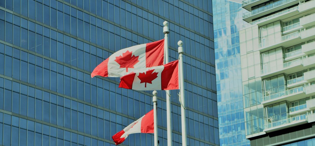 Three Canadian flags waving on tall flagpoles in front of modern glass office buildings with reflections of the sky and surrounding architecture.