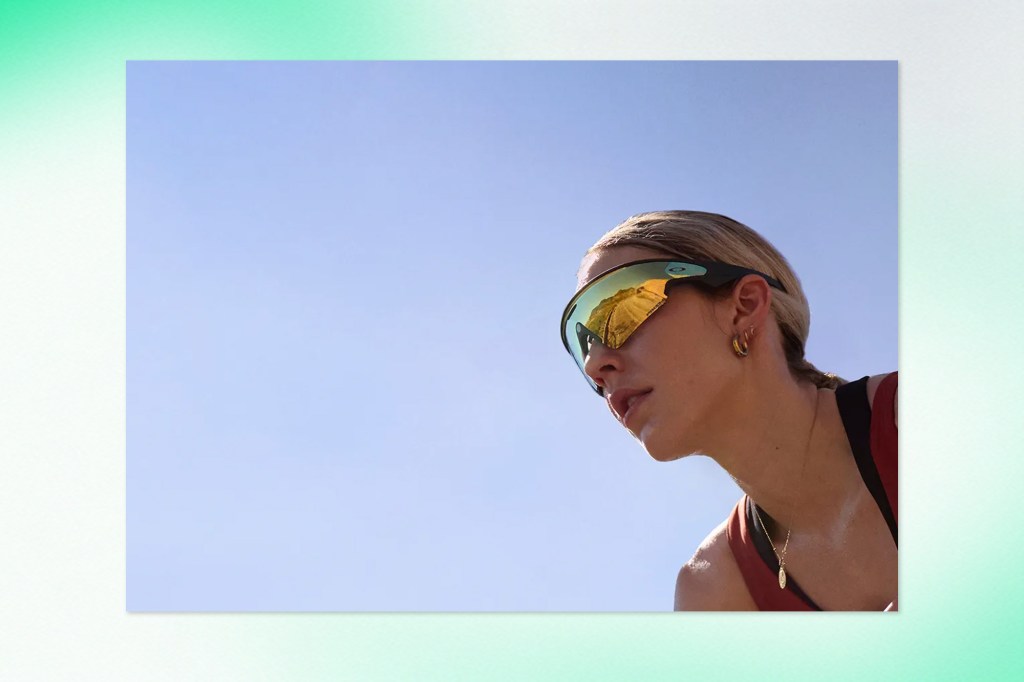 Close-up of a woman wearing sporty Oakley-branded smart glasses with reflective yellow-tinted lenses, looking focused against a clear sky.