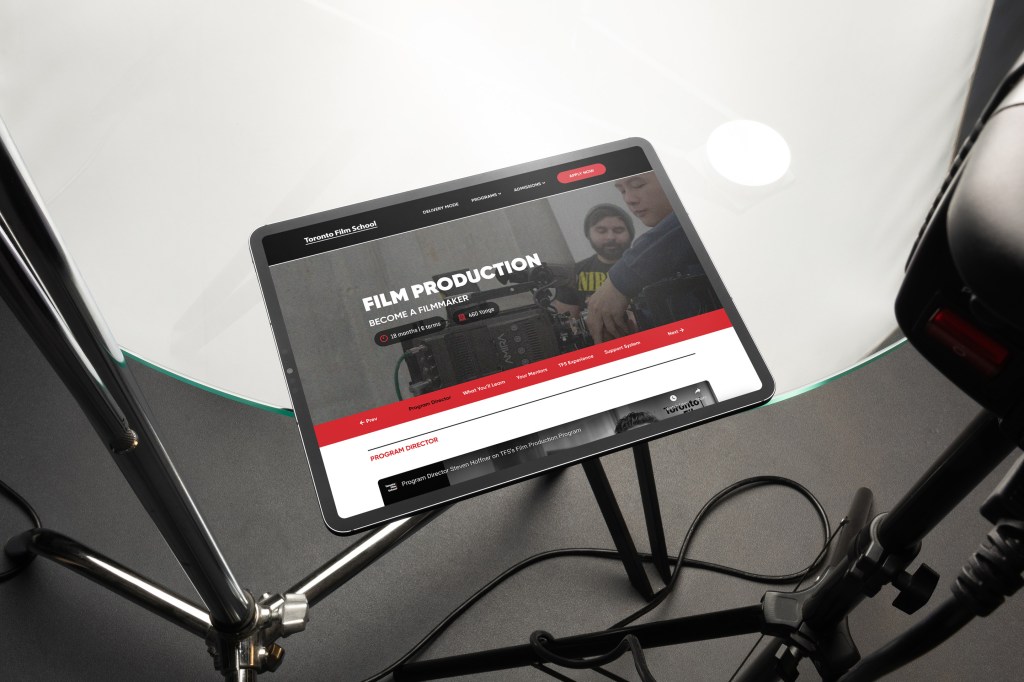 A tablet placed on a tripod stand displays the Toronto Film School website, specifically the Film Production program page. The screen shows the headline “FILM PRODUCTION – BECOME A FILMMAKER” along with navigation tabs and a video of Program Director Steven Hoffner. The setup is positioned over a glass table with surrounding lighting and video equipment, evoking a studio environment.