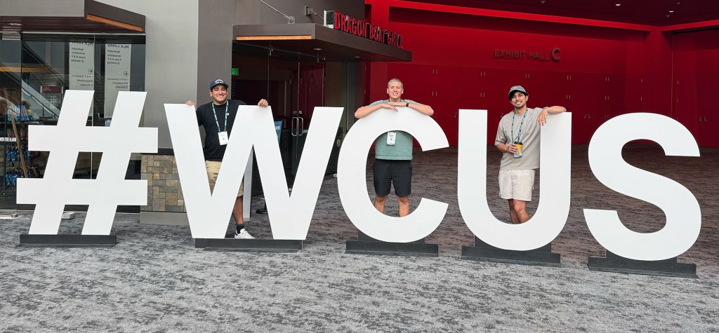 Three members of the Trew Knowledge team stand beside oversized white letters spelling out "#WCUS" inside the Oregon Convention Center.