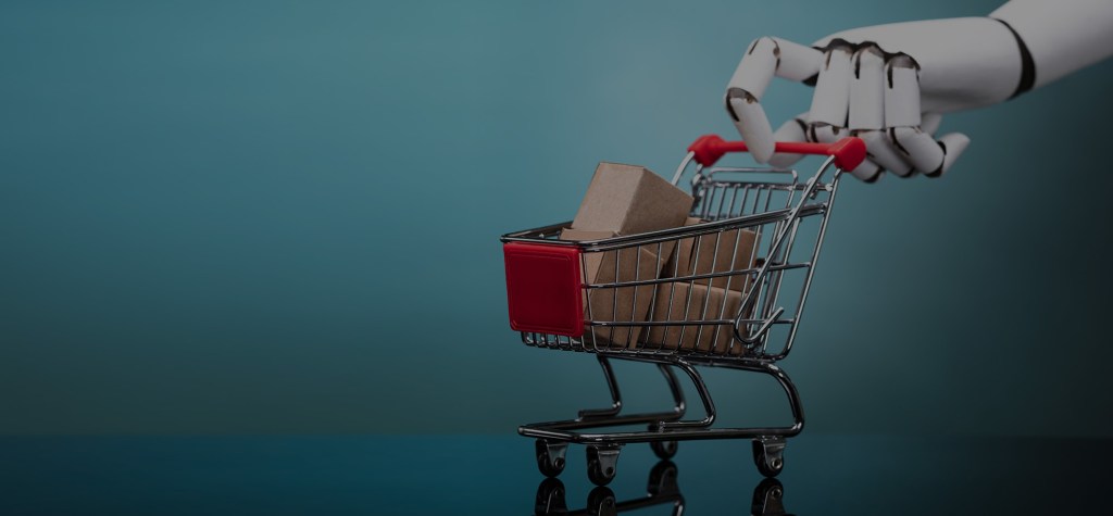 Close-up of a robot hand holding the handle of a miniature shopping cart filled with cardboard boxes, symbolizing AI-driven e-commerce and automation in retail.