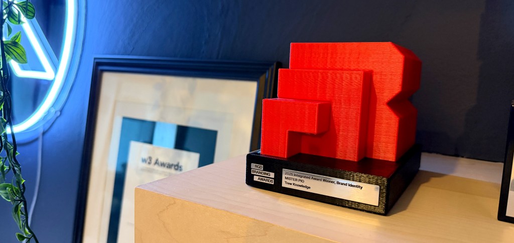 Close-up of the red RGD Branding Award trophy displayed on a wooden shelf. The engraved plaque reads '2025 Integrated Award Winner, Brand Identity – MISTER PIO, Trew Knowledge.' In the background, a framed w3 Awards certificate is partially visible next to a neon sign and trailing plant.