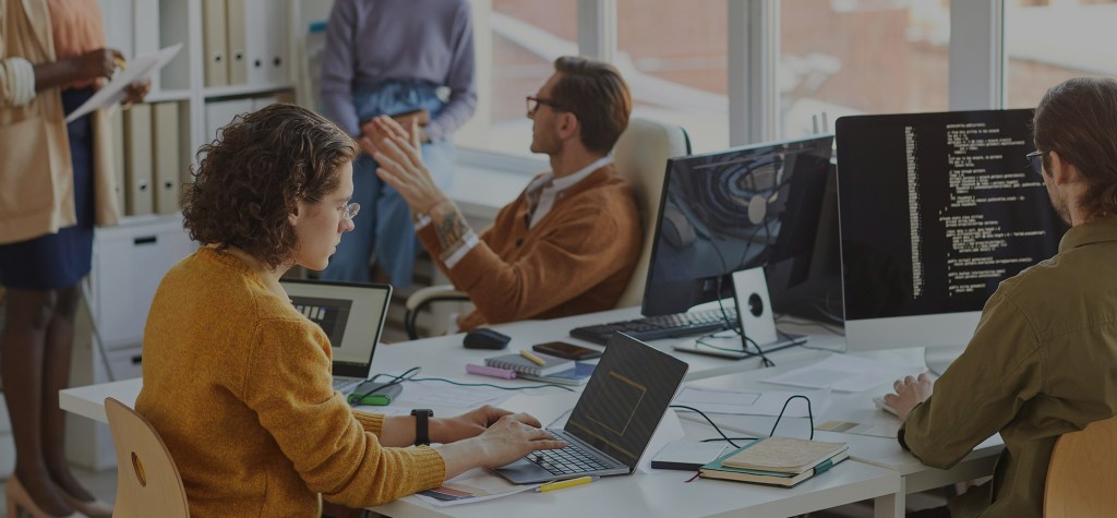 Team members collaborating in a modern office, with one person coding on a desktop and another typing on a laptop.