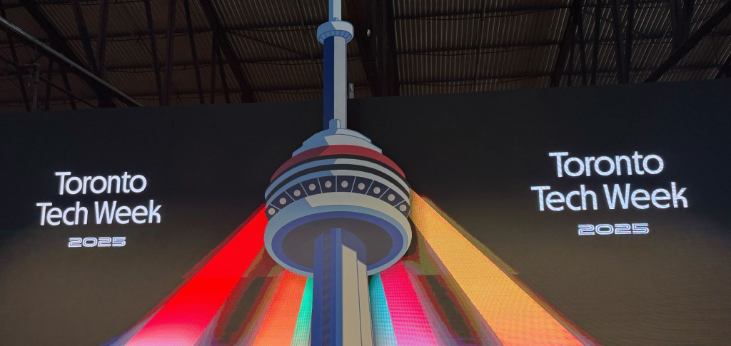 Stage screen at Toronto Tech Week 2025 featuring a digital illustration of the CN Tower lit with vibrant red, orange, and yellow beams, with the text 'Toronto Tech Week 2025' on both sides.