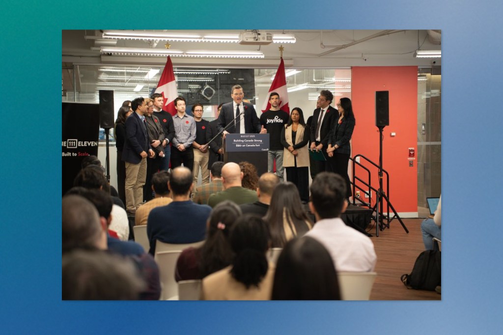 Canada's AI minister, Evan Solomon, speaks at a podium with two Canadian flags behind him. A group of people stands onstage beside him, and an audience watches from seated rows. The backdrop includes branding from OneEleven, a Canadian technology innovation hub.