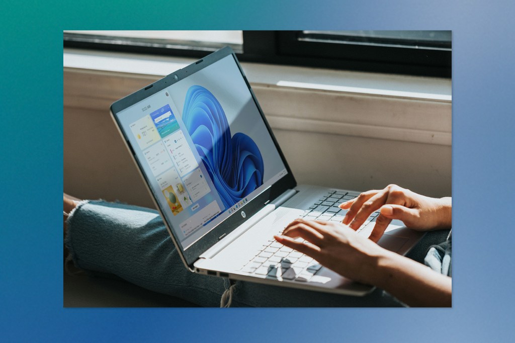 A person typing on a laptop displaying the Windows interface, seated near a bright window.