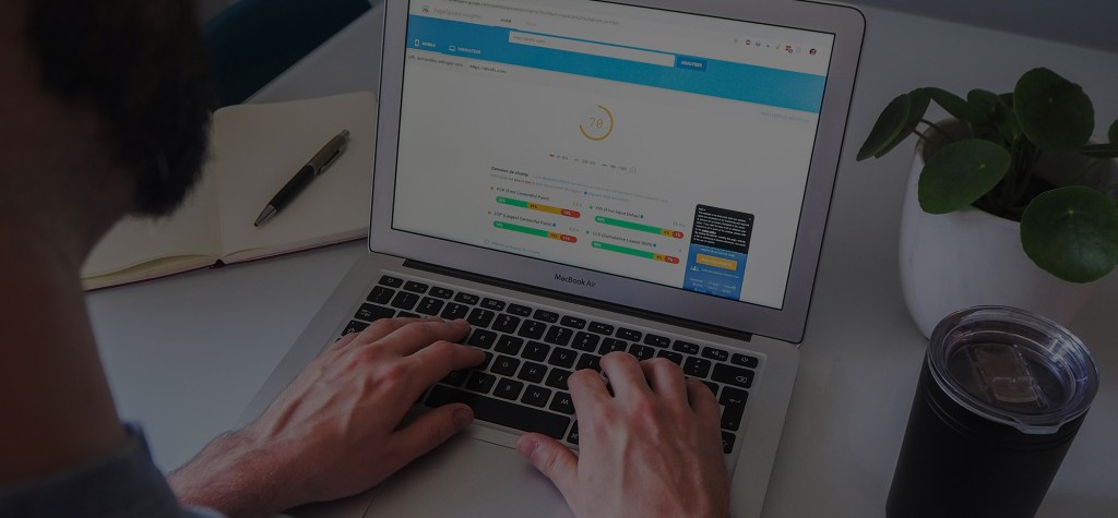 A person working on a MacBook Air, viewing a Google PageSpeed Insights report on the screen. An open notebook, a pen, a plant, and a drink are placed on the desk, creating a workspace setting focused on web performance analysis.