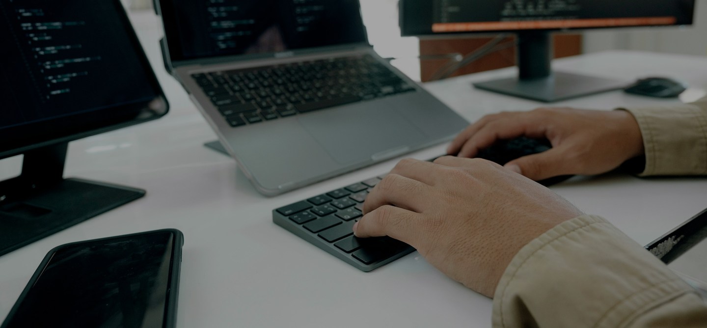 Close-up of a person typing on a compact keyboard at a desk with multiple screens displaying lines of code. A laptop and two monitors show development environments in a dimly lit workspace.
