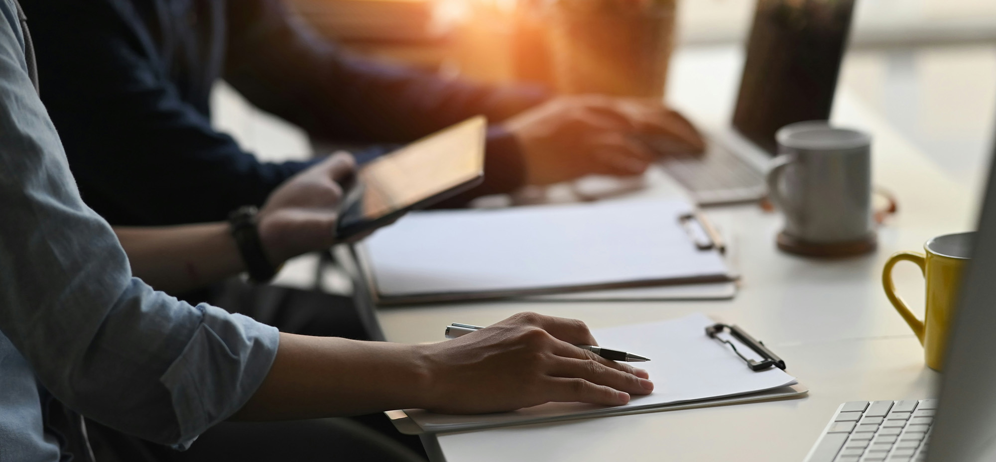 Close-up of two people working at a desk with laptops, clipboards, and coffee mugs in a sunlit office setting.