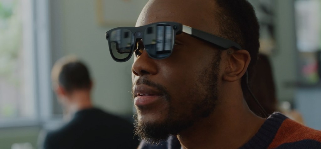 A man wearing dark augmented-reality smart glasses looks ahead in a bright café, with reflections of the room visible on the lenses.