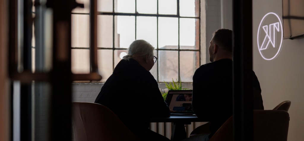 Two people sit at a small round table in a softly lit office, silhouettes outlined against large industrial windows as they look at a laptop screen together. A faint TK logo glows on the wall in the background.