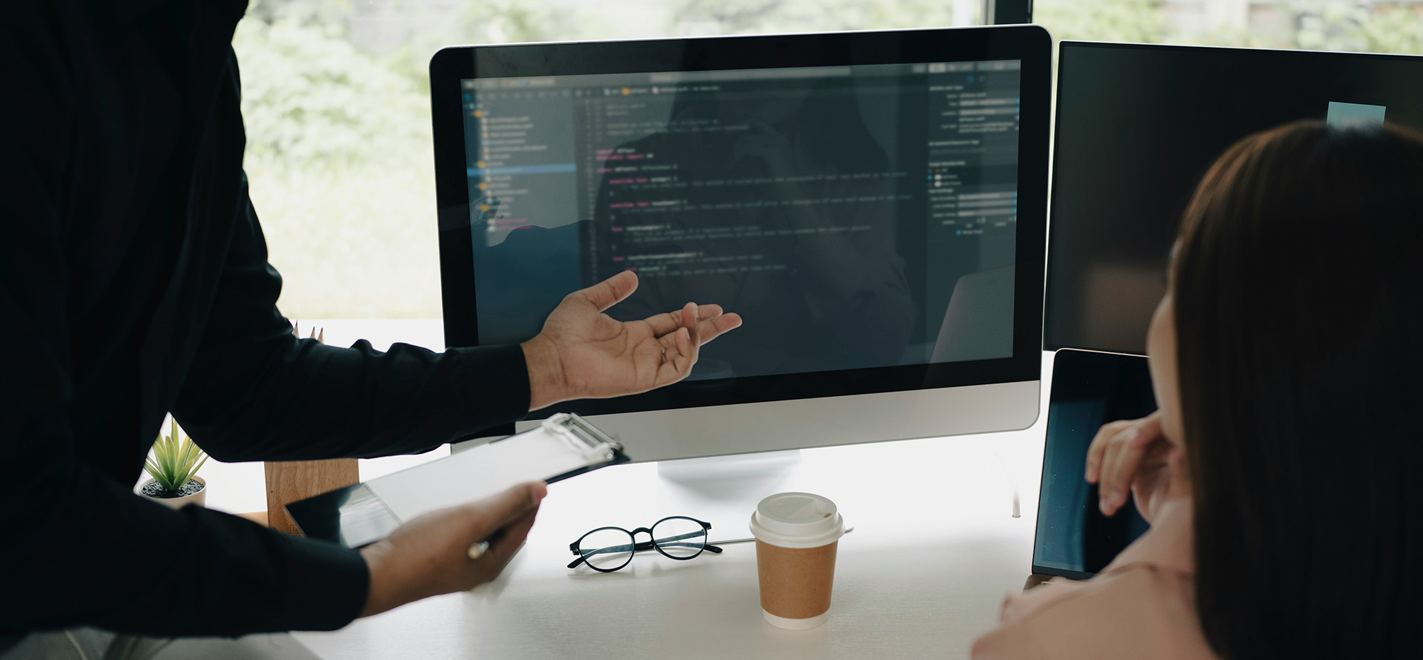 Two people seated at a desk reviewing code displayed on a large desktop monitor, with one person gesturing toward the screen during a technical discussion in a modern workspace.