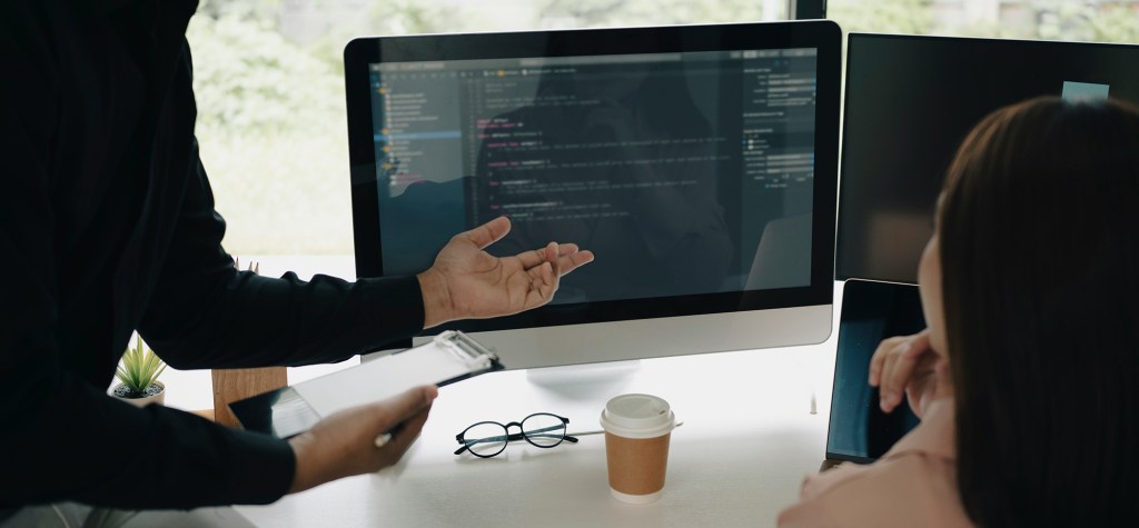 Two people seated at a desk reviewing code displayed on a large desktop monitor, with one person gesturing toward the screen during a technical discussion in a modern workspace.