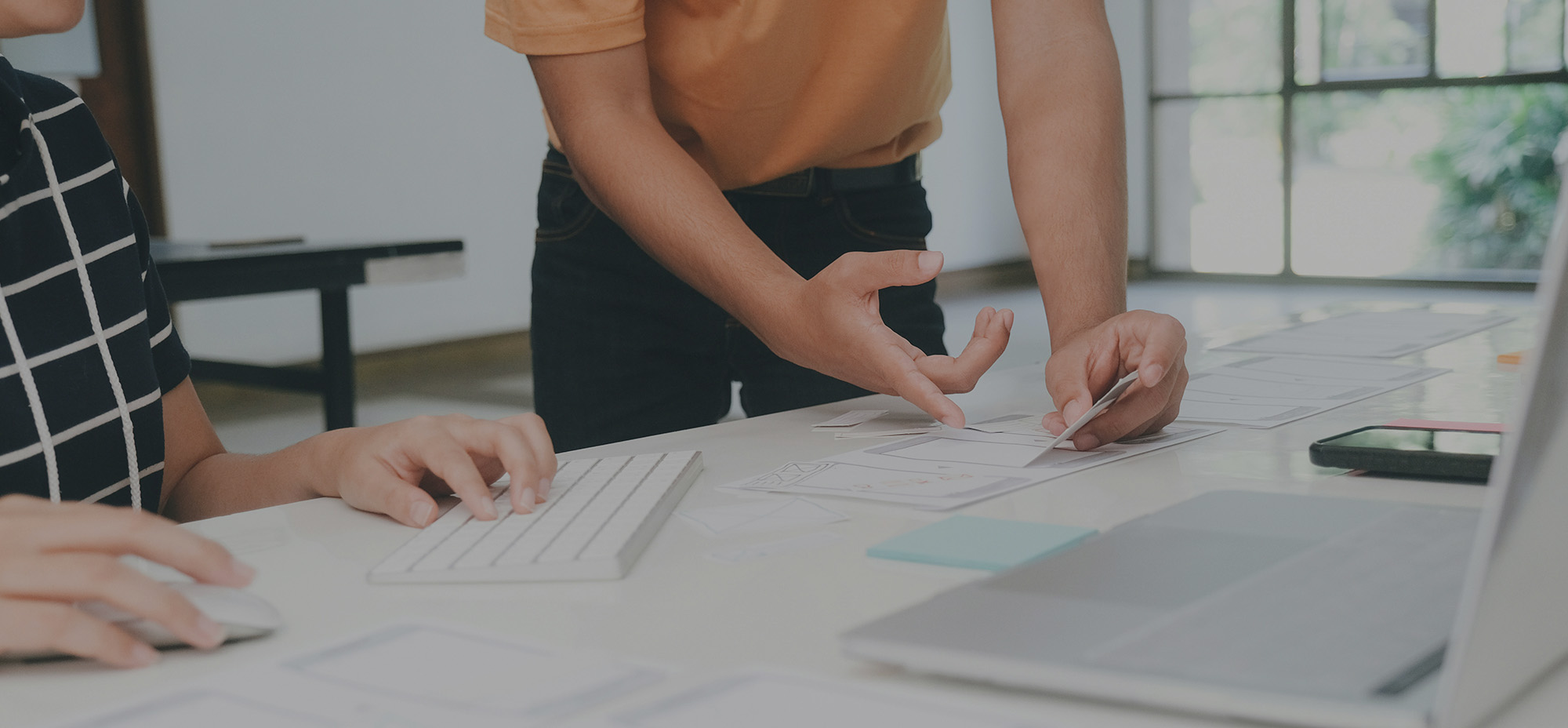 Two people collaborating at a desk, arranging paper wireframes and notes beside a keyboard and laptop, suggesting hands-on interface and user flow design planning.