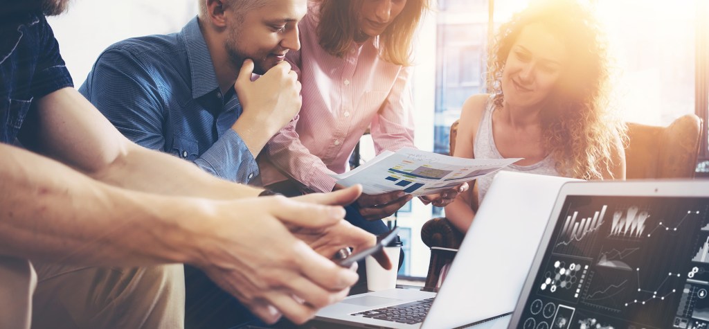 Small group of people collaborating around a laptop, reviewing printed charts and on-screen analytics in a bright office environment.