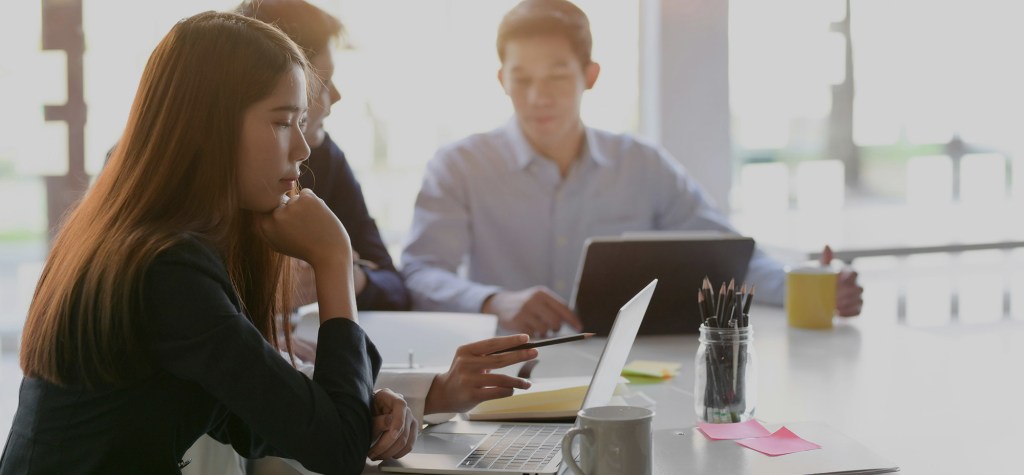 A focused woman sits at a table working on a laptop while holding a pen, with two colleagues slightly out of focus behind her also working on laptops in a bright, modern office setting.