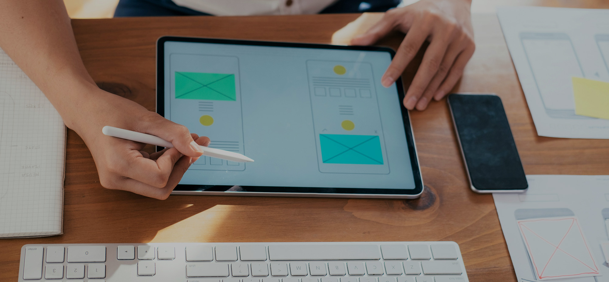 Designer reviewing mobile interface wireframes on a tablet using a stylus, with keyboard, phone, and layout sketches visible on a desk.