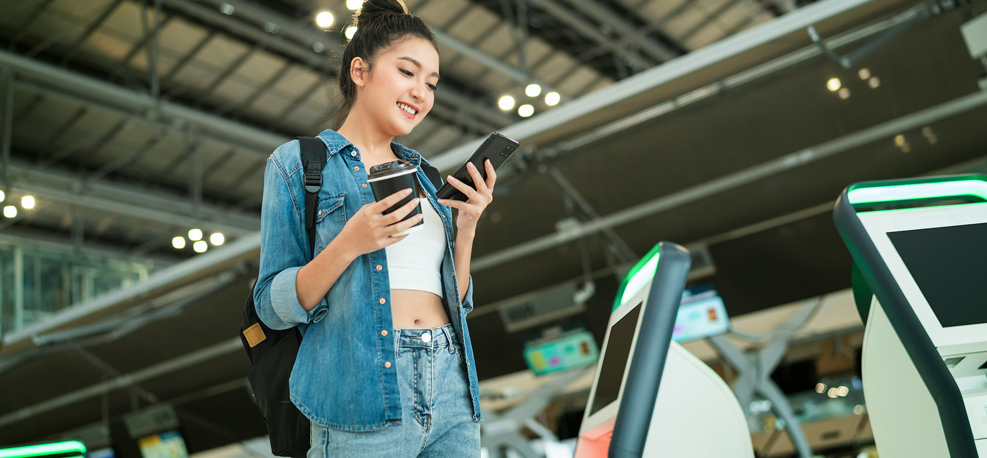 Woman standing in an airport terminal, holding a coffee and checking her smartphone near self-service kiosks, representing on-the-go access to travel information.