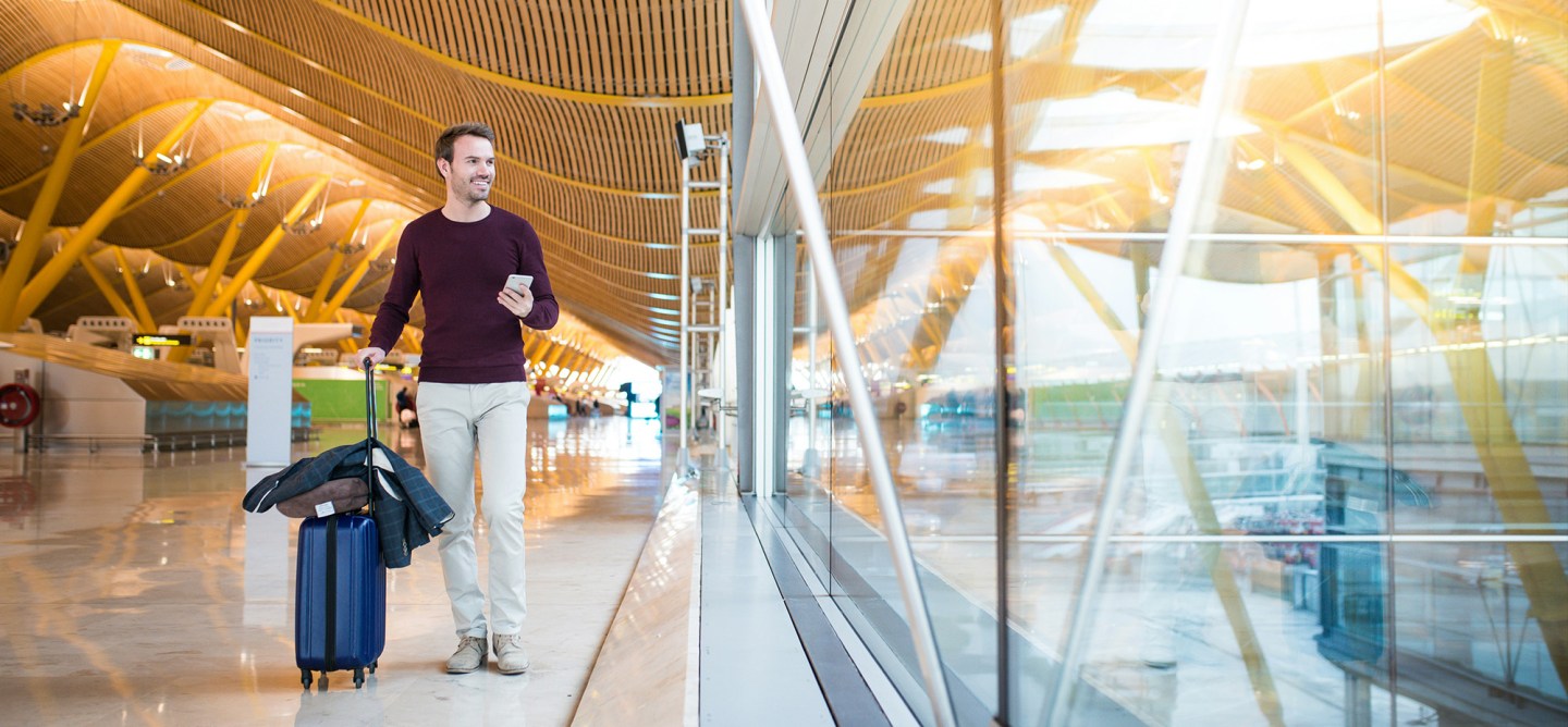 Traveller walking through a modern airport terminal with a rolling suitcase, looking at a smartphone, suggesting real-time updates during a journey.