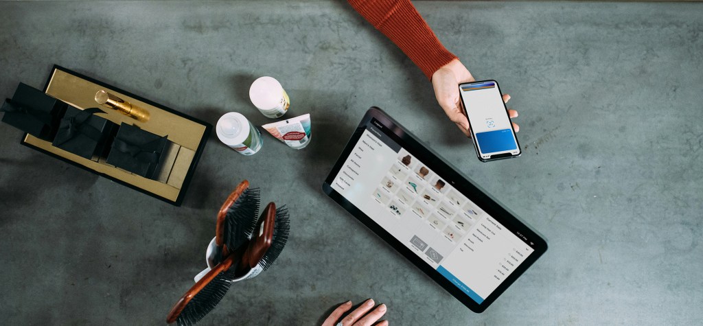 Top-down view of a workspace showing a tablet with a product management or content interface, a smartphone displaying a login or verification screen, and various consumer items on a table, illustrating a connected digital customer journey across devices.